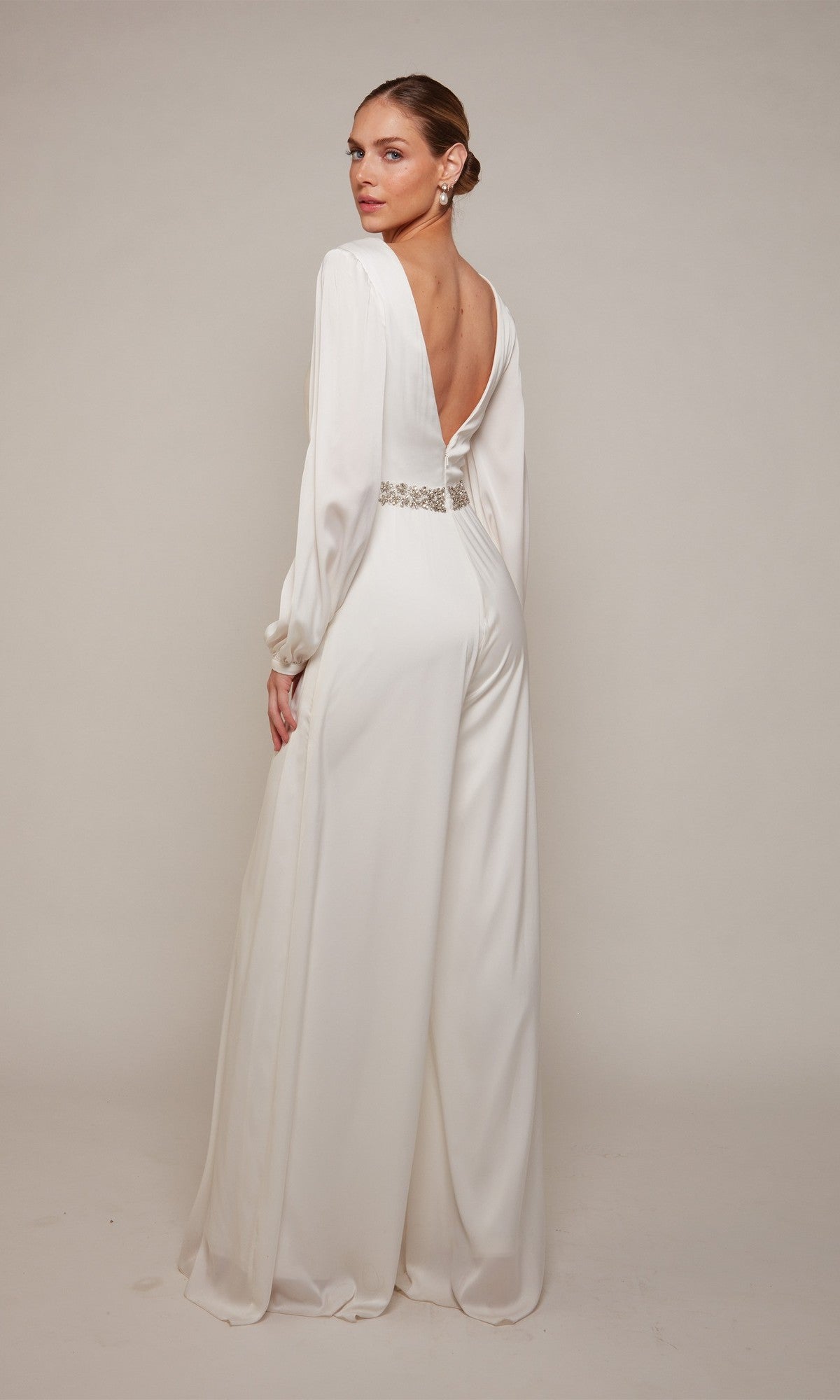 A woman models the Alyce 70031 Long Formal Jumpsuit—a white, floor-length, long-sleeved satin-chiffon style featuring an open back and a decorative waistband—posed against a light background.